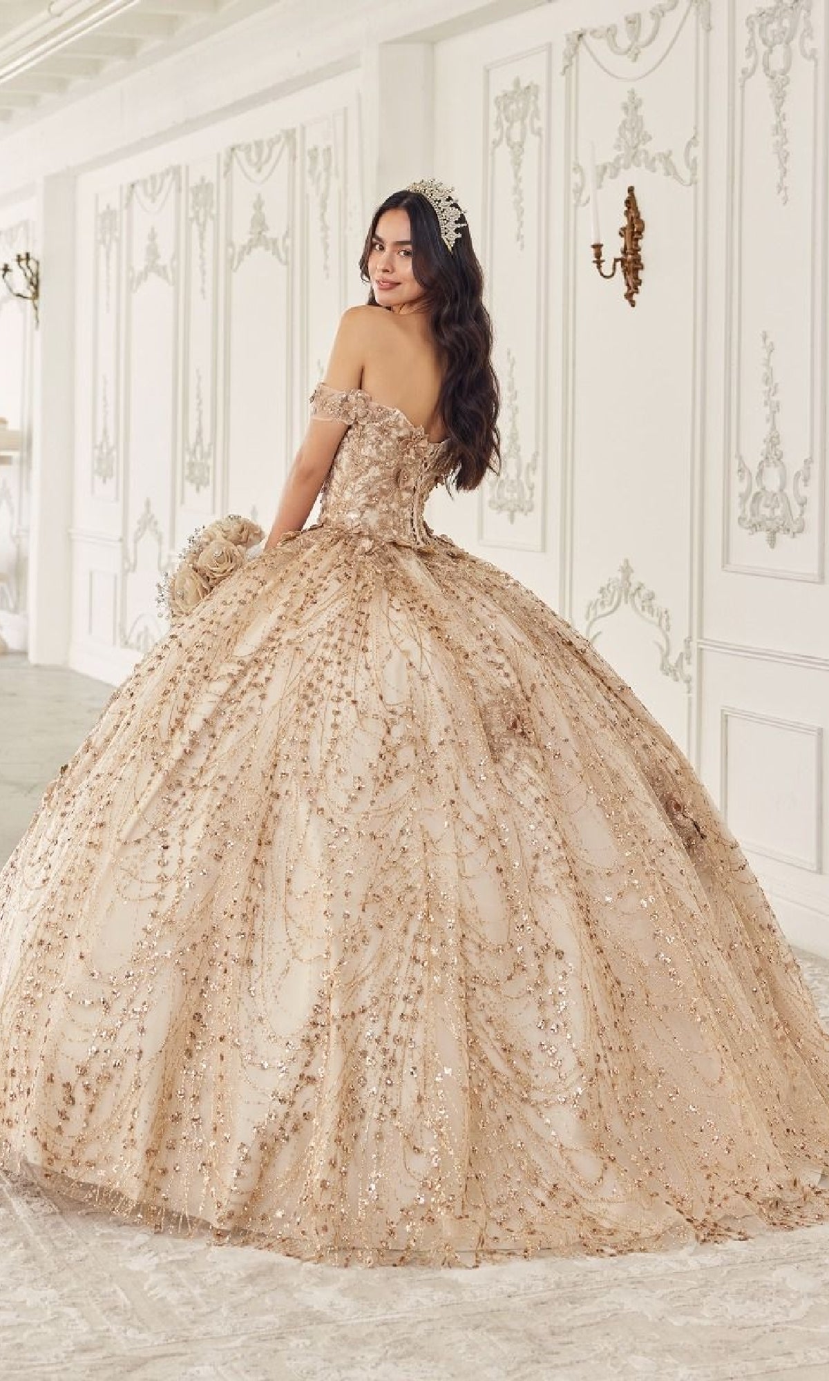 A young woman wearing the Quinceanera Dress 15712 By Ladivine, featuring ornate champagne fabric with intricate beading and delicate floral appliques, stands in an elegant white-decorated room, gazing over her shoulder.